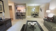 This single-floor residence showcases an open-concept floor plan that seamlessly connects the living, dining, and kitchen areas into one cohesive, functional space. The living area is anchored by a glass-top coffee table set atop a dark area rug, creating a stylish focal point that complements the soft, neutral carpeting extending throughout the main living spaces.

The kitchen is positioned behind a raised breakfast bar, which provides a practical division between the cooking and living areas while maintaining an open sightline. Two bar stools are tucked neatly beneath the counter, offering casual seating. Light wood cabinetry lines the kitchen walls, accompanied by under-cabinet lighting and a white refrigerator, contributing to a clean, functional aesthetic.

Adjacent to the kitchen, the dining area features an intimate round table surrounded by ornate wrought iron chairs. Above, a pendant light fixture with a warm glow draws the eye upward, while a decorative sunburst mirror adorns the wall, adding visual depth. Candle sconces flank the mirror, enhancing the room's classic, welcoming character. Warm beige walls unify the space throughout.