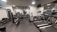 This fitness center is a dedicated, single-floor exercise facility thoughtfully designed to accommodate a full range of workout routines. The space is outfitted with a solid selection of cardio equipment, including treadmills and an elliptical trainer, positioned to allow comfortable use and easy circulation throughout the room. A wall-mounted television is installed near the cardio area, providing entertainment during workouts. The strength training section features a multi-function cable machine, a vertical knee raise and pull-up tower, a flat weight bench, and a collection of free weights and dumbbells, offering users versatile options for resistance training. Full-length mirrors line one wall, visually expanding the space and allowing users to monitor their form during exercise. The flooring is a dark rubberized surface, providing both durability and traction. Overhead pendant lighting and recessed fixtures illuminate the room evenly, creating a focused and functional environment. The clean, neutral color palette of white and grey walls contributes to a modern, professional atmosphere. Overall, this fitness center is a practical and well-organized space suited for a variety of fitness levels and training styles.