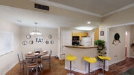 This single-story residence offers a thoughtfully designed layout with interconnected living spaces. The entryway opens into a living area finished with dark hardwood-style flooring and crown molding, with a sliding glass door that draws in natural light and provides access to an outdoor area.

The kitchen is positioned around a peninsula counter topped with granite-style surfaces and features warm wood cabinetry, a full suite of appliances including a refrigerator, microwave, and range, along with a skylight-style ceiling fixture. Vibrant yellow bar stools line the peninsula, adding a pop of color. Adjacent to the kitchen, the dining area accommodates a round wooden table beneath a multi-arm chandelier, complemented by decorative wall accents.

A dedicated laundry room is conveniently accessible directly from the kitchen. The unit includes multiple bedrooms, each equipped with ceiling fans. One bedroom features carpet flooring and an en-suite bathroom, while another offers hardwood-style floors and a walk-in closet. Both bathrooms include warm wood vanities, and one features a shower-tub combination with tile flooring.