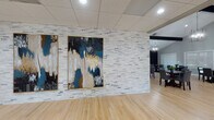 This well-appointed single-floor gathering space combines contemporary design with functional versatility across several distinct areas. A dramatic accent wall clad in stacked light-toned stone panels serves as a focal point, showcasing two large-scale abstract paintings in rich tones of teal, gold, and white framed in gold — setting an artistic, sophisticated tone upon entry.

The main gathering hall features soaring vaulted ceilings with exposed dark wood beams and a geometric chandelier, flooding the space with an open, airy feel. Multiple dining tables with upholstered chairs are arranged throughout, while French doors at the far end invite natural light and provide outdoor access. A spacious lounge area is furnished with plush navy and gray sofas, accent chairs, and a patterned area rug beneath an identical statement chandelier.

A second, more intimate lounge features drop-panel ceilings, recessed lighting, and a curated seating arrangement of neutral accent chairs around a sculptural coffee table, complemented by a navy sideboard and layered artwork.

The full kitchen is outfitted with white shaker cabinetry, granite-look countertops, a mosaic tile backsplash, a beverage refrigerator, and a central island — ideal for catering and events. Light hardwood flooring runs throughout, unifying the space with warmth and continuity.
