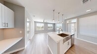 This contemporary single-floor residence showcases a bright, open-concept layout designed for comfortable everyday living. The heart of the home is a well-appointed kitchen featuring crisp white cabinetry, a subway tile backsplash, stainless steel appliances including a refrigerator, range, and microwave, and a generous central island topped with white quartz countertops. A deep farmhouse-style sink with a brushed gold faucet and elegant pendant lighting suspended from chains complete the kitchen's modern aesthetic. Light wood-tone flooring flows seamlessly from the kitchen into the expansive living area, which is bathed in natural light from multiple large windows offering outdoor views. Ceiling fans are present throughout for added comfort. The bedrooms feature plush carpeting, ceiling fans, and windows that frame pleasant outdoor scenery. A spacious walk-in closet with built-in shelving and hanging rods provides ample storage. The bathroom is finished with large-format tile surrounding a soaking tub and shower combination, brushed nickel fixtures, and white cabinetry for additional storage. Neutral gray walls and white trim create a clean, cohesive palette throughout the entire residence.
