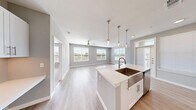 This contemporary single-floor residence showcases a bright, open-concept layout designed for comfortable everyday living. The heart of the home is a well-appointed kitchen featuring crisp white cabinetry, a subway tile backsplash, stainless steel appliances including a refrigerator, range, and microwave, and a generous central island topped with white quartz countertops. A deep farmhouse-style sink with a brushed gold faucet and elegant pendant lighting suspended from chains complete the kitchen's modern aesthetic. Light wood-tone flooring flows seamlessly from the kitchen into the expansive living area, which is bathed in natural light from multiple large windows offering outdoor views. Ceiling fans are present throughout for added comfort. The bedrooms feature plush carpeting, ceiling fans, and windows that frame pleasant outdoor scenery. A spacious walk-in closet with built-in shelving and hanging rods provides ample storage. The bathroom is finished with large-format tile surrounding a soaking tub and shower combination, brushed nickel fixtures, and white cabinetry for additional storage. Neutral gray walls and white trim create a clean, cohesive palette throughout the entire residence.