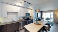 This single-floor space showcases a thoughtfully designed open-concept layout where the kitchen and living area connect seamlessly. The kitchen is a standout feature, equipped with a full suite of stainless steel appliances, including a range with oven, built-in microwave, and dishwasher. Crisp white upper cabinetry contrasts elegantly with dark lower cabinets, while dark stone countertops add a sophisticated touch. A white waterfall-edge kitchen island provides additional workspace and casual seating with bar-height chairs, anchored above by a metallic drum pendant light. Track lighting runs the length of the kitchen ceiling, ensuring ample task illumination. Light wood-style flooring extends throughout, creating visual continuity between spaces. The adjacent living area features a bold navy accent wall that adds depth and contrast, complemented by neutral furnishings and a floor lamp. A large window allows natural light to fill the living space, enhancing the bright, airy feel. The overall aesthetic is contemporary and cohesive, blending functional design with modern style.