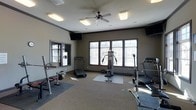 This fitness center offers a comprehensive workout environment designed to accommodate a variety of exercise routines. The space is anchored by large windows that allow abundant natural light to fill the room, creating a bright and energizing atmosphere. The neutral-toned walls and carpeted flooring contribute to a clean, no-frills aesthetic focused on functionality.

The room is outfitted with a well-rounded selection of equipment, including multiple treadmills for cardio training, a multi-station cable machine for resistance and strength exercises, and a free weights area complete with a barbell rack and adjustable bench. Protective rubber mats are positioned beneath the equipment, ensuring safety and floor preservation.

Overhead, a ceiling fan provides air circulation throughout the space, complemented by flush-mounted rectangular light fixtures that ensure consistent illumination. Wall-mounted flat-screen televisions are positioned at multiple vantage points, allowing users to stay entertained during their workouts. A large wall mirror on one side of the room enhances the sense of space and allows for proper form checking during exercises. Overall, this fitness center delivers a practical and comfortable environment suitable for a full range of training needs.