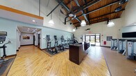 This fitness center is designed to accommodate a comprehensive workout experience within a single, open-plan floor. The space immediately impresses with its soaring ceilings, which feature exposed wooden beams, industrial-style ductwork, and large pendant light fixtures that illuminate the room with a warm, inviting glow. A ceiling fan adds to the comfort of the workout environment.

The main exercise area is outfitted with polished hardwood flooring across a generous open space, transitioning to rubberized flooring beneath the equipment zones. The cardio section lines one wall and includes multiple treadmills, elliptical trainers, and a stationary bike, each equipped with individual media screens. A central dark-wood station with built-in amenities serves as a functional hub within the space.

The strength and functional training area is richly equipped with cable machines, a weight rack stocked with dumbbells, exercise balls, benches, and a wall-mounted climbing ladder. Full-length mirrors span multiple walls, enhancing the sense of openness and allowing users to monitor their form throughout their sessions. A flat-screen television is mounted for added convenience.

Adjacent to the main floor, a tiled corridor leads to restroom and shower facilities, finished with decorative mosaic tile accents and dark-toned floor tiles, providing a clean and practical transition space from the gym floor.