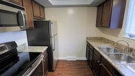 This single-floor space includes a well-appointed kitchen that serves as a central feature. The kitchen is designed with a galley-style layout, maximizing efficiency through its parallel arrangement of cabinetry and countertops. Rich, dark-stained wood cabinetry lines both sides of the kitchen, providing ample storage with upper and lower cabinet configurations. The countertops are finished in light granite, offering a pleasing contrast against the darker cabinetry. Stainless steel appliances, including a refrigerator, electric range, and dishwasher, contribute a modern and cohesive aesthetic. The sink is positioned along one counter run, complementing the overall functional design. Underfoot, dark hardwood flooring adds warmth and continuity throughout the space. Neutral, light-toned walls keep the kitchen feeling open and bright. A floor-level vent indicates a forced-air HVAC system, and a recessed ceiling detail adds a subtle architectural element overhead. The overall ambiance of the kitchen is practical yet polished, combining classic dark wood tones with modern finishes for a timeless and functional environment.