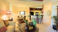 This single-floor residence features an open-concept layout that seamlessly connects the living, dining, and kitchen areas into one cohesive, functional space. The living area is anchored by a plush armchair and a decorative trunk-style coffee table atop a mosaic area rug, creating an inviting focal point. Lush tropical houseplants add a vibrant, natural element throughout.

The dining area sits adjacent, furnished with dark wood chairs beneath a classic chandelier, while a mirrored wall sconce arrangement adds visual depth. A warm yellow-toned color palette flows throughout, complemented by bold terracotta-red accent walls that define transitions between spaces, adding character and dimension.

The kitchen is highlighted by cream cabinetry, black appliances, and a raised breakfast bar with tall dark wood barstools — ideal for casual dining. A skylight-style overhead fixture brightens the kitchen area, enhancing the overall warmth of the space. Crown molding and light-toned carpeting contribute a polished, cohesive finish. The overall ambiance is cozy, colorful, and well-appointed, with thoughtful decorative details throughout.