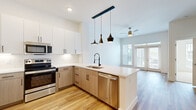 This single-story unit offers a thoughtfully designed open-concept layout that maximizes its efficient footprint. The combined kitchen and living area serves as the heart of the space, featuring light hardwood flooring, elevated ceilings, and a set of French doors flanked by floor-to-ceiling windows that draw in abundant natural light and provide outdoor access. A ceiling fan complements the airy atmosphere of the living area.

The kitchen is outfitted with a full suite of stainless steel appliances, including a refrigerator, electric range, built-in microwave, and dishwasher. Cabinetry combines crisp white upper cabinets with warm wood-toned lower cabinets, creating a contemporary two-tone aesthetic. A subway tile backsplash, light quartz countertops, and a peninsula with a matte black industrial-style faucet and undermount sink complete the functional and stylish cooking space. Pendant lighting in matte black and gold tones hangs above the peninsula, adding a striking design detail.

The bedroom is a calm, neutrally finished room with plush carpeting, white walls, a ceiling fan, and a window fitted with blinds. A dedicated closet provides convenient storage. Throughout the unit, clean lines and a consistent neutral palette create a cohesive, modern aesthetic.