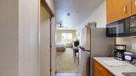 Located at 225 112th Ave NE in Bellevue, Washington, this studio unit offers a smartly organized single-floor layout that maximizes every square foot. The main living and sleeping area features a patterned carpet, a ceiling fan for year-round comfort, and a window that brings in natural light. The bedroom is furnished with a king-sized bed, flanking nightstands with lamps, a wall-mounted flat-screen TV, a multi-drawer dresser, and a dedicated work desk with a chair — making the space functional for both rest and productivity. A cozy armchair with a side table adds a relaxed seating option, while an open closet with a curtain provides accessible storage. The kitchenette, positioned near the entrance, is equipped with a full-sized stainless steel refrigerator, wood-toned cabinetry, and a countertop workspace. The bathroom features a tub-shower combination with a curtain, a wall-mounted sink, a toilet, a mirrored medicine cabinet, and stone-look tile flooring, offering a clean and practical design. The unit's neutral color palette and warm lighting throughout contribute to a calm and comfortable ambiance, well-suited for extended stays in the heart of Bellevue.
