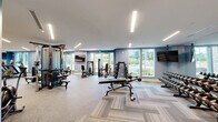 This modern fitness center in Huntersville, NC offers a spacious and thoughtfully designed environment for a full range of workout activities. The facility spans a generous footprint across two dedicated exercise rooms, connected by a hallway and complemented by a bathroom and sauna.

The primary exercise room is bright and open, featuring large windows that allow abundant natural light and views of the surrounding greenery. It is outfitted with a robust selection of strength training equipment, including barbells, weight plates, a power rack, cable machines, and a variety of adjustable benches. Floor-to-ceiling mirrors line the walls, enhancing the sense of space and aiding proper form. The room's flooring features a distinctive herringbone-patterned carpet tile that adds visual interest throughout.

The secondary exercise room is dedicated to cardio and cycling, housing multiple treadmills, ellipticals, stair climbers, and stationary bikes. Wall-mounted flat-screen TVs provide entertainment, while large windows keep the space airy and connected to the outdoors.

The hallway connects the exercise areas and includes a dual water fountain and bottle-filling station set against stylish stone-look tile walls. The facility also features a beautifully crafted cedar sauna with tiered benches and warm ambient lighting, providing an ideal space for post-workout recovery. Linear LED lighting throughout the facility creates a clean, energizing atmosphere.