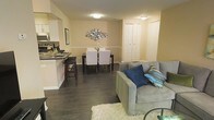 This single-floor residence offers an open-concept layout that integrates the living, dining, and kitchen areas into one cohesive and functional space. The interior is finished with rich, dark hardwood flooring that runs throughout, providing a warm and grounded aesthetic that contrasts beautifully with the soft, neutral-toned walls accented by classic white chair rail molding.

The living area is anchored by a comfortable sofa adorned with decorative throw pillows, complemented by a plush white area rug that adds texture and warmth underfoot. Adjacent to the living space, the dining area features a dark wood dining table paired with white upholstered chairs, creating an elegant contrast. A decorative metallic wall sculpture serves as a focal point, adding a touch of artistic character to the space.

The kitchen flows seamlessly from the dining area, separated by a breakfast bar with dark stone countertops and bar seating — ideal for casual dining or entertaining. The kitchen is equipped with modern appliances and ample cabinetry. Flush-mount ceiling lights provide consistent, even illumination throughout the open space. A set of bi-fold closet doors offers convenient built-in storage. Overall, the space presents a clean, modern, and inviting atmosphere well-suited for comfortable everyday living.