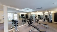 This fitness center offers a thoroughly equipped workout environment designed to support a wide range of exercise routines. The space is finished with neutral carpeting and features a smooth white ceiling with recessed light panels and HVAC venting, providing consistent, even illumination throughout. Large mirrors line the walls, visually expanding the room while allowing users to monitor their form during workouts. A wall-mounted flat-screen television adds a functional amenity for entertainment during training sessions. The strength training area is well-stocked, featuring multiple tiers of dumbbells on organized racks, covering a broad range of weights to accommodate all fitness levels. Weight benches, including adjustable and flat variants from Life Fitness, are positioned throughout the floor space for versatile use. Cable and multi-station machines are arranged along the perimeter, offering additional resistance training options. Windows along one wall allow natural light to filter into the space, complementing the artificial lighting and contributing to an open, energizing atmosphere. The overall layout is thoughtfully organized, balancing equipment density with sufficient open floor space to allow for comfortable movement between stations.