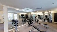 This fitness center offers a thoroughly equipped workout environment designed to support a wide range of exercise routines. The space is finished with neutral carpeting and features a smooth white ceiling with recessed light panels and HVAC venting, providing consistent, even illumination throughout. Large mirrors line the walls, visually expanding the room while allowing users to monitor their form during workouts. A wall-mounted flat-screen television adds a functional amenity for entertainment during training sessions. The strength training area is well-stocked, featuring multiple tiers of dumbbells on organized racks, covering a broad range of weights to accommodate all fitness levels. Weight benches, including adjustable and flat variants from Life Fitness, are positioned throughout the floor space for versatile use. Cable and multi-station machines are arranged along the perimeter, offering additional resistance training options. Windows along one wall allow natural light to filter into the space, complementing the artificial lighting and contributing to an open, energizing atmosphere. The overall layout is thoughtfully organized, balancing equipment density with sufficient open floor space to allow for comfortable movement between stations.