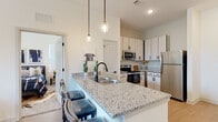 This well-appointed single-floor residence offers a thoughtfully designed open-concept layout that seamlessly connects the kitchen, dining, and living areas. The kitchen is a standout feature, showcasing light wood cabinetry with matte black hardware, granite countertops, and a full suite of stainless steel appliances including a refrigerator, range, microwave, and dishwasher. A large center island with a double sink and pendant lighting above serves as both a functional workspace and a casual dining area with bar seating. Light wood-style flooring flows throughout the main living areas, enhancing the bright and airy feel created by multiple large windows. The living room benefits from generous natural light and views of lush greenery outside. The bedroom features plush carpeting and a sizable window that continues the theme of natural light. The space includes two bathrooms: one featuring a granite-topped vanity with ample storage and a shower-tub combination, and a secondary half bath with a matching vanity and light wood flooring. The overall design maintains a clean, neutral palette with modern finishes, creating a cohesive and contemporary living environment throughout.