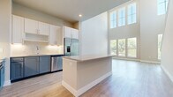 This well-designed three-story home distributes its substantial living area thoughtfully across three floors. The entry level serves as a welcoming base, while the main living floor above hosts the combined kitchen and living room — a bright, open-concept space highlighted by soaring double-height windows that flood the area with natural light and offer outdoor views. The kitchen features white upper cabinetry paired with slate-blue lower cabinets, stainless steel appliances, a white tile backsplash, a central island with quartz-style countertops, and light hardwood flooring that extends into the living area. Two staircases with dark wood treads and white risers connect all three levels. The uppermost floor contains all three bedrooms, four bathrooms, and three closets, including a spacious primary bedroom with balcony access, a ceiling fan, and plush carpeting. Secondary bedrooms also feature carpet, ceiling fans, and built-in closets. Bathrooms are finished with large-format neutral tiles, blue-gray vanities, and soaking tub-shower combinations. Walk-in closets include built-in shelving and hanging rods. Neutral tones and clean lines define the overall aesthetic throughout.