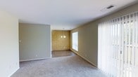 This single-floor home offers a comfortable and functional layout with a warm, understated aesthetic. The living area is finished with plush carpet flooring and painted in neutral tones, creating a calm and inviting atmosphere. It flows seamlessly into the dining area, which is distinguished by a striking yellow brick accent wall that adds character and a touch of vintage charm to the space. The dining area features hardwood-style flooring, providing a clean contrast to the carpeted living zone, and is illuminated by a pendant chandelier that adds a refined focal point overhead. Large sliding glass doors in the dining area invite an abundance of natural light and offer views of the outdoors, enhancing the overall sense of openness. Additional windows fitted with vertical blinds further contribute to the bright, airy feel of the space. White trim and baseboards throughout provide a crisp, polished finish that ties the rooms together cohesively. The single-floor layout ensures ease of movement and accessibility, making this a practical yet characterful space well-suited for everyday living and entertaining.