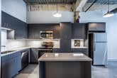 This one-bedroom, one-bathroom unit in Memphis, Tennessee showcases an appealing blend of industrial architecture and contemporary design. The space retains striking original elements throughout, including exposed ceiling structures with visible beams, ductwork, and aged materials that lend an authentic loft character to the interior.

The kitchen is a standout feature, outfitted with slate-gray flat-front cabinetry extending to the ceiling for ample storage, granite-look countertops, a white tile backsplash, and a full suite of stainless steel appliances including a range, microwave, and refrigerator. A central island provides additional prep and seating space, while polished concrete floors extend throughout the main living areas, enhancing the industrial aesthetic.

The bedroom benefits from natural light through large windows framed by exposed brick walls, adding warmth and texture to the space. A ceiling fan contributes to comfort. The bathroom is clean and well-appointed, featuring a granite-topped vanity with dark cabinetry, a full tub-shower combination, and neutral tile flooring. The overall ambiance of the unit is modern and urban, with thoughtful finishes that complement its historic industrial framework.