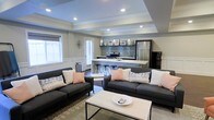 This single-floor space showcases a thoughtfully designed open-concept layout that seamlessly connects the living and kitchen areas into one cohesive, functional environment. The living area is anchored by two charcoal gray sofas accented with blush and patterned throw pillows, arranged around a modern white and black coffee table, creating an inviting and contemporary seating arrangement. Natural light filters in through multiple windows dressed with shutters and blinds, lending a bright and airy feel to the space. A defining architectural feature is the tray ceiling with recessed lighting, which adds depth and a refined character to the room. The kitchen is well-appointed with stainless steel appliances, including a range with a tile backsplash surround and a full-size refrigerator. Pendant lights hang gracefully above a white breakfast bar with industrial-style bar stools, serving as both a casual dining spot and a visual divider between the kitchen and living zones. Neutral gray walls and dark hardwood flooring provide a sophisticated backdrop that ties the design together, while subtle decorative touches throughout contribute to a polished, modern aesthetic. The open flow and cohesive design make this space feel both spacious and welcoming.