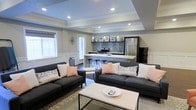 This single-floor space showcases a thoughtfully designed open-concept layout that seamlessly connects the living and kitchen areas into one cohesive, functional environment. The living area is anchored by two charcoal gray sofas accented with blush and patterned throw pillows, arranged around a modern white and black coffee table, creating an inviting and contemporary seating arrangement. Natural light filters in through multiple windows dressed with shutters and blinds, lending a bright and airy feel to the space. A defining architectural feature is the tray ceiling with recessed lighting, which adds depth and a refined character to the room. The kitchen is well-appointed with stainless steel appliances, including a range with a tile backsplash surround and a full-size refrigerator. Pendant lights hang gracefully above a white breakfast bar with industrial-style bar stools, serving as both a casual dining spot and a visual divider between the kitchen and living zones. Neutral gray walls and dark hardwood flooring provide a sophisticated backdrop that ties the design together, while subtle decorative touches throughout contribute to a polished, modern aesthetic. The open flow and cohesive design make this space feel both spacious and welcoming.