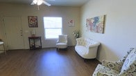 This single-floor space offers a warm and inviting atmosphere, thoughtfully designed to balance comfort and style. The living area features rich, dark hardwood-style flooring that extends throughout, providing a polished foundation that complements the neutral, softly toned walls. Natural light filters in through a large window, brightening the room and creating an airy, open feel. A ceiling fan overhead adds both functionality and a classic touch to the space.

The seating area is furnished with elegant tufted pieces, including a loveseat and a coordinating accent chair, both upholstered in crisp white fabric that adds a refined, sophisticated character to the room. Decorative wall art in vibrant, floral-inspired patterns provides colorful focal points, lending personality and visual interest to the otherwise understated palette. A wooden side table and a small console table with a lamp contribute to the layered, homey ambiance.

Overall, the space strikes a balance between casual comfort and tasteful décor, making it well-suited for relaxed conversation or quiet leisure. The cohesive design elements — from the warm flooring to the carefully chosen furnishings and artwork — come together to create a space that feels both functional and aesthetically curated.