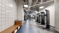 Located at 1235 Martin St in Nashville, TN, this two-story fitness facility offers a comprehensive athletic environment spanning two generously sized floors. The ground floor serves as the primary fitness space, featuring a large open gym area with polished terrazzo-style flooring in contrasting tones. This level houses an extensive free weights section with dumbbell racks, benches, and multi-functional cable machines, complemented by a turf strip styled like a football field for functional training. Large floor-to-ceiling windows flood the space with natural light, while exposed ceilings with linear LED lighting and visible ductwork contribute an industrial aesthetic. A bold mural and warm wood accents near the staircase add visual character. The upper floor is anchored by a full-sized basketball court featuring a hardwood floor, wall-mounted basketball hoops, padded safety wall panels, and vibrant large-scale graffiti-style mural art. Oversized windows along one wall provide views of the surrounding Nashville skyline. Additional rooms include dedicated cardio areas equipped with ellipticals and stationary bikes, a multipurpose studio with a Reflect mirror and punching bag, and connecting corridors linking all zones seamlessly throughout both levels.