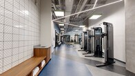 Located at 1235 Martin St in Nashville, TN, this two-story fitness facility offers a comprehensive athletic environment spanning two generously sized floors. The ground floor serves as the primary fitness space, featuring a large open gym area with polished terrazzo-style flooring in contrasting tones. This level houses an extensive free weights section with dumbbell racks, benches, and multi-functional cable machines, complemented by a turf strip styled like a football field for functional training. Large floor-to-ceiling windows flood the space with natural light, while exposed ceilings with linear LED lighting and visible ductwork contribute an industrial aesthetic. A bold mural and warm wood accents near the staircase add visual character. The upper floor is anchored by a full-sized basketball court featuring a hardwood floor, wall-mounted basketball hoops, padded safety wall panels, and vibrant large-scale graffiti-style mural art. Oversized windows along one wall provide views of the surrounding Nashville skyline. Additional rooms include dedicated cardio areas equipped with ellipticals and stationary bikes, a multipurpose studio with a Reflect mirror and punching bag, and connecting corridors linking all zones seamlessly throughout both levels.
