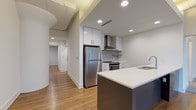 Located on Euclid Avenue in Cleveland's vibrant midtown corridor, this single-floor unit masterfully combines historic character with modern design. The space is immediately distinguished by its stunning arched, leaded-glass windows, which flood the living and bedroom areas with abundant natural light while offering an elegant architectural nod to the building's storied past. Exposed beams along the ceiling further enhance the sense of history and craftsmanship throughout.

The kitchen is a standout feature, outfitted with stainless steel appliances including a full-size refrigerator and range with a wall-mounted hood vent, white upper cabinetry, and a boldly accented navy-blue island with a white quartz countertop and undermount double sink. Warm hardwood flooring runs throughout the main living areas, creating a cohesive and inviting atmosphere.

The bedroom features plush carpeting and benefits from the same striking arched windows, providing a bright, airy retreat. The bathrooms are finished with large-format light tile flooring, floating vanities with dark cabinetry, and a tub-shower combination with tasteful tile surround. A second bathroom offers a walk-in tiled shower. Recessed lighting and clean white walls complete the contemporary, move-in-ready aesthetic of this well-appointed space.