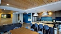 This single-floor space showcases a thoughtfully designed open-concept layout that brings together a fully equipped kitchen, dining area, and lounge in one cohesive environment. The kitchen is outfitted with stainless steel appliances, including a full-size refrigerator and microwave, dark wood cabinetry, and a granite-topped island that doubles as a casual dining bar. Large dome pendant lights in a brushed silver finish hang above the island, providing both functional illumination and a bold design statement. The dining area features a generous wood-toned table surrounded by sleek blue upholstered chairs, complementing the space's signature color palette. Bold blue accent walls punctuate the interior, adding depth and vibrancy alongside neutral white and grey tones. A wood-paneled ceiling adds warmth and architectural interest overhead, contrasting beautifully with the polished concrete flooring below. The lounge area is furnished with a dark sectional sofa and coordinating accent pieces, creating a comfortable retreat within the larger open floor plan. Abstract wall art echoes the blue tones throughout, tying the aesthetic together. The overall ambiance is modern and inviting, balancing professional functionality with a welcoming, stylish atmosphere.