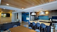 This single-floor space showcases a thoughtfully designed open-concept layout that brings together a fully equipped kitchen, dining area, and lounge in one cohesive environment. The kitchen is outfitted with stainless steel appliances, including a full-size refrigerator and microwave, dark wood cabinetry, and a granite-topped island that doubles as a casual dining bar. Large dome pendant lights in a brushed silver finish hang above the island, providing both functional illumination and a bold design statement. The dining area features a generous wood-toned table surrounded by sleek blue upholstered chairs, complementing the space's signature color palette. Bold blue accent walls punctuate the interior, adding depth and vibrancy alongside neutral white and grey tones. A wood-paneled ceiling adds warmth and architectural interest overhead, contrasting beautifully with the polished concrete flooring below. The lounge area is furnished with a dark sectional sofa and coordinating accent pieces, creating a comfortable retreat within the larger open floor plan. Abstract wall art echoes the blue tones throughout, tying the aesthetic together. The overall ambiance is modern and inviting, balancing professional functionality with a welcoming, stylish atmosphere.