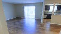 This single-floor space offers a clean, functional layout designed for comfortable everyday living. The main living area is generously proportioned, showcasing rich hardwood-style flooring that runs throughout and reflects natural light beautifully. A large sliding glass door fitted with vertical blinds serves as the focal point of the living space, flooding the room with abundant natural light and providing direct access to an outdoor area with views of surrounding greenery.

The open-concept design allows the living area to flow seamlessly into the kitchen, which is positioned just off to the side. The kitchen features dark espresso-toned upper cabinetry paired with light stone-look countertops, creating a pleasant contrast. A dishwasher is visible among the appliances, hinting at the practical amenities included.

The walls are finished in a neutral warm beige tone that complements both the flooring and cabinetry, contributing to a cohesive and inviting atmosphere throughout. Textured ceilings add a subtle architectural detail overhead. The overall ambiance of the space is bright, airy, and move-in ready, with a straightforward layout that lends itself well to a variety of furnishing arrangements and personal styles.