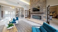 This single-floor community clubhouse is thoughtfully designed to offer both comfort and functionality across a series of well-connected spaces. The centerpiece of the main lounge is a dramatic floor-to-ceiling stacked stone fireplace with a built-in linear hearth and wall-mounted television above a solid wood mantel, creating a striking focal point. Soaring ceilings with crown molding and an abundance of large windows and French doors flood the interior with natural light, while offering expansive views of the outdoor pool and landscaped grounds. Stylish seating arrangements with patterned upholstered chairs and teal velvet sofas contribute to a sophisticated yet welcoming ambiance. Light wood-look tile flooring flows throughout, complementing the neutral color palette. A modern kitchenette features floor-to-ceiling dark espresso cabinetry, stone countertops, a mosaic tile backsplash, and a stainless steel refrigerator. A cozy lounge nook includes built-in banquette seating with a round table, ideal for informal gatherings. The dedicated office area is outfitted with a built-in desk, open shelving with pendant lighting, and ample storage drawers. The main entry corridor features soaring ceilings, structural columns, and dark wood framed glass partitions that define spaces with architectural elegance.