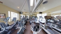 This fitness center is housed within a single-story space distinguished by its vaulted ceiling with prominent exposed white beams that lend a sense of openness and architectural character. Skylights integrated into the roofline allow natural light to pour in, complementing the large windows lining the walls, which further brighten the interior and create an airy, inviting atmosphere.

The facility is outfitted with a comprehensive selection of cardio equipment, including treadmills, elliptical trainers, stationary upright and recumbent bikes, and stair climbers, arranged to maximize usable floor space while maintaining comfortable circulation paths between machines. Ceiling fans are installed throughout to aid in air circulation during workouts, and mounted flat-screen televisions provide entertainment options for users. A full-length mirror along one wall visually expands the space and allows members to monitor their form. The flooring features a durable, dark rubberized surface appropriate for high-traffic fitness use. Overall, the space is functional, well-lit, and thoughtfully designed to support a well-rounded cardio workout experience.