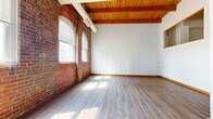 Located at 1300 West 9th Street in Cleveland's vibrant Ohio City neighborhood, this two-floor residence masterfully blends industrial heritage with contemporary design. The expansive main floor features soaring ceilings with warm wooden beam work and exposed brick walls that lend authentic loft character throughout. Wide-plank hardwood flooring flows through the open living areas, while natural light pours in through arched and oversized windows. The kitchen is well-appointed with white shaker cabinetry, a classic subway tile backsplash, stainless steel appliances, and hardwood floors — offering both style and functionality. Hallways lined with exposed brick and wooden accents connect the spaces seamlessly. The three bedrooms are generously proportioned; the primary bedroom features plush carpeting, multiple windows with urban views, and direct access to an outdoor deck. Additional bedrooms include high windows framing brick architectural details and ample closet space with built-in wire shelving systems. The two bathrooms are cleanly finished with tile flooring, full tub-shower combinations, and wide vanity mirrors. The upper floor offers a more intimate footprint, ideal for bedrooms or private retreats. Throughout, the interplay of raw brick, natural wood, and clean white walls creates a warm, urban ambiance.