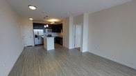 This single-floor residence offers a thoughtfully designed open-concept layout that seamlessly connects the living and kitchen areas, creating a spacious and airy feel. The kitchen is a standout feature, showcasing dark espresso cabinetry that contrasts beautifully against light-toned walls and neutral hardwood-style flooring that extends throughout the space. Stainless steel appliances — including a French door refrigerator with an ice dispenser, a microwave, a range, and a dishwasher — provide a polished, contemporary finish. A central kitchen island with a light stone countertop offers additional prep space and serves as a natural gathering point. Pendant lighting hangs above the island, adding warmth and a refined touch to the kitchen's aesthetic. The open living area is generously proportioned, providing ample room for flexible furniture arrangements. Neutral walls and consistent flooring unify the space, while recessed lighting ensures the interiors remain bright and welcoming. A white paneled door is visible toward the rear of the space, hinting at additional rooms beyond. The overall ambiance is clean, modern, and move-in ready, suited for those who appreciate functional design with a contemporary edge.