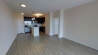 This single-floor residence offers a thoughtfully designed open-concept layout that seamlessly connects the living and kitchen areas, creating a spacious and airy feel. The kitchen is a standout feature, showcasing dark espresso cabinetry that contrasts beautifully against light-toned walls and neutral hardwood-style flooring that extends throughout the space. Stainless steel appliances — including a French door refrigerator with an ice dispenser, a microwave, a range, and a dishwasher — provide a polished, contemporary finish. A central kitchen island with a light stone countertop offers additional prep space and serves as a natural gathering point. Pendant lighting hangs above the island, adding warmth and a refined touch to the kitchen's aesthetic. The open living area is generously proportioned, providing ample room for flexible furniture arrangements. Neutral walls and consistent flooring unify the space, while recessed lighting ensures the interiors remain bright and welcoming. A white paneled door is visible toward the rear of the space, hinting at additional rooms beyond. The overall ambiance is clean, modern, and move-in ready, suited for those who appreciate functional design with a contemporary edge.