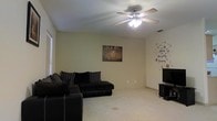 This single-floor residence offers a straightforward and functional layout centered around a cozy living area. The main living space is finished with soft carpet flooring that extends throughout, providing a warm and comfortable underfoot feel. A ceiling fan with an integrated light fixture is mounted overhead, offering both ambient lighting and efficient air circulation. The room is furnished with a dark sectional sofa, making it an ideal space for relaxation and gathering. Natural light enters through a window fitted with horizontal blinds, brightening the neutral-toned walls. Decorative touches, including a framed wall piece and a playful star-themed wall decal, add a personalized character to the space. A media console positioned in the corner of the room provides a dedicated area for entertainment. The overall ambiance is casual and inviting, with a clean and open feel that lends itself well to comfortable, everyday living. The single-floor design ensures easy accessibility and a cohesive flow throughout the home.