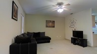 This single-floor residence offers a straightforward and functional layout centered around a cozy living area. The main living space is finished with soft carpet flooring that extends throughout, providing a warm and comfortable underfoot feel. A ceiling fan with an integrated light fixture is mounted overhead, offering both ambient lighting and efficient air circulation. The room is furnished with a dark sectional sofa, making it an ideal space for relaxation and gathering. Natural light enters through a window fitted with horizontal blinds, brightening the neutral-toned walls. Decorative touches, including a framed wall piece and a playful star-themed wall decal, add a personalized character to the space. A media console positioned in the corner of the room provides a dedicated area for entertainment. The overall ambiance is casual and inviting, with a clean and open feel that lends itself well to comfortable, everyday living. The single-floor design ensures easy accessibility and a cohesive flow throughout the home.
