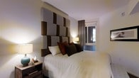 This single-floor residence showcases a carefully curated interior design with a strong emphasis on comfort and sophistication. The bedroom serves as a standout feature, anchored by a dramatic floor-to-ceiling upholstered headboard composed of geometric panels in alternating shades of taupe, grey, and deep brown — creating a bold visual focal point. The bed is dressed in crisp white linens and layered with richly toned accent pillows in burnt orange, chocolate brown, and ivory, adding warmth and depth to the room. Soft ambient lighting from bedside table lamps enhances the cozy, intimate atmosphere, complementing the room's neutral wall tones. A window dressed with tailored curtains in a warm grey hue allows for natural light while maintaining privacy, offering a glimpse of greenery beyond. The nightstands are finished in dark wood, contributing to the room's grounded, earthy palette. The space overall reflects a refined, modern aesthetic with a cohesive blend of textures and tones that create an inviting and restful environment throughout.