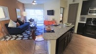 This single-floor residence is designed with an open-concept layout that seamlessly connects the kitchen, dining, and living areas into one cohesive, functional space. The kitchen features a prominent granite-topped island with bar seating, offering both a prep surface and a casual dining spot. Dark cabinetry beneath the island provides ample storage and creates a striking contrast against the light granite surface. Hardwood-style flooring runs throughout, adding warmth and continuity across the space.

The living area is bright and welcoming, benefiting from large sliding glass doors with vertical blinds that allow abundant natural light to filter in. A ceiling fan overhead contributes to the airy feel of the room. The open sightlines from the kitchen through to the living space make the area feel expansive and well-connected.

A glimpse into the adjacent bedroom reveals a continuation of the thoughtful layout, accessible through a nearby doorway. The overall ambiance is modern and well-appointed, with clean lines, contemporary finishes, and a neutral color palette accented by bold furnishing choices. This space is well-suited for both everyday living and casual entertaining.