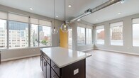 Located at 150 N 3rd St in the heart of downtown Columbus, Ohio, this modern single-floor unit offers a stylish and well-appointed living experience with striking urban views. The open-concept living and kitchen area is anchored by a large central island with a crisp white countertop and dark cabinetry, illuminated by globe pendant lights that add a contemporary touch. Exposed ductwork lends an industrial character to the space, while light wood-tone flooring runs throughout, creating a warm, cohesive aesthetic. The kitchen is thoughtfully designed with dark lower cabinets, a white subway tile backsplash, open wood shelving, stainless steel appliances including a dishwasher and range, and a matte black faucet. Large windows flood the living area with natural light and frame impressive views of the Columbus skyline, complemented by a bold yellow entry door that serves as a vibrant design accent. The bedroom is a calm, neutral space with hardwood-style flooring and a window providing natural light. The bathroom features a light blue vanity with a white countertop, a sleek framed mirror, matte black fixtures, and a glass-enclosed walk-in shower with clean tile surround. This unit combines modern design with an unbeatable downtown location.