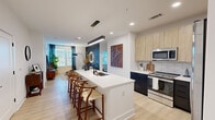 This thoughtfully designed single-floor residence offers 1 bedroom, 2 bathrooms, a dedicated office, and a walk-in closet. The open-concept kitchen and living area serves as the heart of the home, anchored by a large waterfall-edge island with a deep undermount sink and seating for multiple bar stools. The kitchen features a two-tone cabinet design — light wood uppers paired with dark lower cabinets — complemented by a geometric tile backsplash and a full suite of stainless steel appliances, including a French door refrigerator, range, and built-in microwave. A sleek linear pendant light defines the island space. The living room is highlighted by a bold dark accent wall and large windows that invite natural light. The bedroom offers a comfortable layout with a ceiling fan, natural light from dual windows, and direct access to a well-appointed en-suite bathroom. The bathroom features a double-sink vanity with matte black fixtures, dark cabinetry, and leads directly into a spacious walk-in closet with built-in shelving. The office is a bright, versatile room with a ceiling fan and generous window views. Light hardwood-style flooring flows consistently throughout, unifying the clean, modern aesthetic of the entire space.
