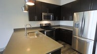 This single-floor space showcases a thoughtfully designed kitchen that serves as a central highlight. The kitchen features a bold, contemporary aesthetic defined by dark espresso-toned cabinetry that extends to the upper and lower levels, providing generous storage throughout. Sleek silver bar-pull hardware adds a refined, modern touch to the cabinetry design.

The countertops are finished in a smooth, neutral-toned surface that complements the dark cabinetry and offers ample workspace. A peninsula-style island extends the counter space further, complete with an undermount single-basin sink and a modern arc-neck faucet, making it ideal for both food preparation and casual use.

Stainless steel appliances, including an electric range, over-the-range microwave, and refrigerator, are integrated seamlessly into the layout, reinforcing the kitchen's contemporary character. The flooring features dark wood-tone planks that ground the space and complement the overall color palette. Pendant lighting overhead adds warmth and dimension. The layout is efficiently organized in a U-shape, optimizing workflow and functionality throughout the cooking space.