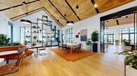 Located on Valley View in East Hanover, NJ, this single-floor amenity space offers a generously sized, open-plan interior defined by striking architectural character. The defining feature is the dramatic vaulted ceiling clad in warm-toned wood planks, supported by bold exposed beams and anchored by a series of statement pendant lights that illuminate the space throughout. Wide-plank hardwood flooring runs continuously across the entire floor, lending a cohesive and inviting feel.

The main gathering area is divided into distinct functional zones. A welcoming lounge near the entrance features plush, richly colored sofas and accent chairs arranged around a sleek linear fireplace set against a white brick accent wall. Floor-to-ceiling black-framed windows and glass doors flood the interior with natural light and provide views to the surrounding outdoor areas.

A central dining and meeting zone is anchored by a large wooden table with black chairs, set atop a vibrant orange patterned rug. Adjacent to this, an open-plan kitchenette with dark cabinetry and modern appliances supports social gatherings. A curved banquette seating area lined with lush indoor plants adds a relaxed, organic touch, while a decorative open shelving unit serves as a bold room divider. The space is thoughtfully curated with artwork and abundant greenery throughout.