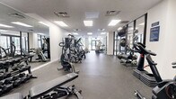 Located at 330 W First St in downtown Dayton, Ohio, this fitness center offers a well-equipped, single-floor exercise space designed to support a full range of workout routines. The room is generously proportioned, providing ample open floor space for movement and equipment layout without feeling crowded.

The facility features a diverse selection of exercise equipment, including multiple treadmills, elliptical trainers, a rowing machine, stationary and air bikes, cable weight machines, a multi-functional strength station, and a weight bench with free weights. Rubber speckled flooring throughout provides durability, shock absorption, and traction during workouts.

Natural light is a defining feature of this space. Large floor-to-ceiling windows and glass-paneled double doors line the perimeter, offering views of the surrounding downtown streetscape and allowing daylight to fill the room. Full-length mirrors are strategically positioned along the walls, visually expanding the space and supporting proper form during exercises.

Overhead, recessed flat-panel lighting supplements the natural light, ensuring consistent illumination throughout all areas of the room. The textured ceiling and clean white walls contribute to a bright, modern aesthetic. The space connects visually to an adjacent area through glass partitions, adding to the open and airy atmosphere of the overall fitness environment.