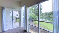 This single-floor space includes a sunroom or enclosed porch area that serves as a standout architectural feature. Large floor-to-ceiling windows line the exterior wall, flooding the space with natural light while framing an unobstructed view of a tranquil waterway surrounded by mature trees and manicured greenery. The screened enclosure allows fresh air to circulate while maintaining a comfortable interior environment. Neutral-toned carpeting underfoot complements the light, airy atmosphere created by the white window frames and pale walls. A sliding glass door connects the interior directly to an outdoor deck with metal railing, extending the living space outdoors and enhancing the connection to the natural surroundings. The deck overlooks a landscaped yard that leads to the water's edge. The overall ambiance is calm and nature-focused, making this sunroom an ideal space for relaxation. The seamless blend of indoor comfort and outdoor scenery is a defining characteristic of this space.