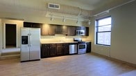 This single-floor space showcases an industrial-modern aesthetic defined by exposed cylindrical ductwork running along the ceiling, light hardwood flooring, and an open layout that creates a sense of spaciousness throughout. The main living area is bathed in natural light streaming through multiple large, black-framed windows that offer views of the surrounding urban streetscape. Track lighting mounted to the ceiling provides additional illumination, complementing a pendant light fixture in one corner of the room. Structural columns are subtly integrated into the open floor plan without disrupting the flow of the space.

The kitchen is designed as a galley-style setup along one wall, featuring rich, dark wood cabinetry paired with light stone-look countertops. Stainless steel appliances — including a refrigerator, gas range, built-in microwave, and dishwasher — provide a polished, contemporary look. A window adjacent to the kitchen extends natural light into this functional area. The exposed ductwork continues throughout, maintaining a cohesive industrial character. Overall, the space combines raw architectural elements with modern finishes to create a practical yet stylish urban environment.