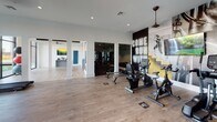 This fitness center is thoughtfully designed across a single floor, offering distinct workout zones that cater to a wide range of exercise needs. The space is divided into several functional areas, each with its own character and equipment selection.

The cardio zone features treadmills, ellipticals, a stair climber, and stationary bikes, including spin-style cycles. Bold navy accent walls and large-scale motivational wall murals—including a prominent "Come Run With Us" graphic—energize the space. Light hardwood-style flooring runs throughout the main areas, complemented by recessed lighting and pendant-style ceiling fans.

Floor-to-ceiling black-framed windows flood a dedicated stretching and functional training area with natural light, offering views of an outdoor courtyard. This section houses a rack of exercise balls and connects to a mirrored yoga and flexibility studio equipped with storage shelving, yoga mats, and a wall-mounted television.

The strength training zone transitions to darker rubber flooring and features floor-to-ceiling mirrors that visually expand the room. Free weights, kettlebells, adjustable benches, and cable machines are well-organized throughout. Neon accent lighting adds a modern, motivating atmosphere to this area.