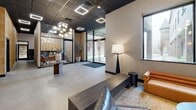 Located at 18 Lane Ave in Caldwell, NJ, this single-floor commercial space offers a thoughtfully designed lobby and reception environment that blends modern aesthetics with functional layout. The entrance immediately draws the eye with a striking feature wall clad in warm, earthy stone tiles, flanked by elevator doors with dramatic marble-style stone surrounds. Polished concrete floors extend throughout, providing a seamless and sophisticated base that ties the space together.

The open lobby area is furnished with a curated lounge arrangement, including cognac leather seating, a round coffee table atop a woven area rug, and a statement floor lamp, creating a welcoming yet professional atmosphere. A sculptural globe chandelier and recessed square LED lighting fixtures illuminate the dark ceiling, adding a bold contrast that enhances the contemporary feel. Large windows allow natural light to filter in, offering views of the surrounding brick exterior.

A glass-enclosed private office sits prominently within the space, featuring warm wood cabinetry, a conference table with leather chairs, and pendant globe lighting — ideal for meetings or private consultations. A dedicated lounge nook with open shelving, curated décor, and additional seating rounds out the space, offering a relaxed yet refined area for visitors and occupants alike.