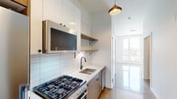 This single-floor space showcases a thoughtfully designed contemporary interior with a cohesive modern aesthetic throughout. The kitchen is a standout feature, equipped with a stainless steel gas range, built-in microwave, and dishwasher. White upper cabinetry is paired with warm wood-toned lower cabinets, creating a balanced two-tone design. A white quartz countertop extends across the workspace, complemented by a stainless steel undermount sink with a matte black faucet. Horizontal subway tile lines the backsplash, adding clean texture, while open wood shelving with black bracket hardware provides functional display space. A pendant light with a brass and black finish hangs above, adding a refined accent. The adjacent living area features polished concrete floors that reflect light, contributing to an open and airy atmosphere. Tall ceilings enhance the sense of volume throughout. A large window at the end of the space draws in abundant natural light and offers exterior views. The bathroom maintains the same modern palette, featuring a white floating vanity with black hardware, a recessed medicine cabinet mirror, and clean white walls, delivering a minimalist and functional finish.