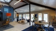This single-story space showcases a striking open-plan layout defined by soaring vaulted ceilings with white shiplap paneling and bold dark-stained exposed structural beams. Linear pendant lighting runs along the ceiling lines, providing both functional illumination and a modern industrial accent. The interior is divided into two primary zones: a lounge area furnished with navy upholstered sofas, armchairs, and cushioned ottomans arranged atop a patterned area rug, and a dining area featuring a rustic round wooden table surrounded by cross-back chairs on a deep blue rug. Light hardwood flooring flows throughout, contrasting with the navy and neutral color palette on the walls. Large windows along multiple walls flood the interior with natural light and offer views of an outdoor area that appears to include a pergola and pool. A bar-height counter with stools is visible along one wall, adding a casual social element. Decorative touches such as wall art, potted plants, and accent lighting contribute to a polished yet relaxed ambiance, making the space well-suited for gathering and socializing.
